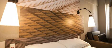 A hotel room with a bed, two bedside lamps, a headboard with a textured pattern, a chair, and a wall socket.