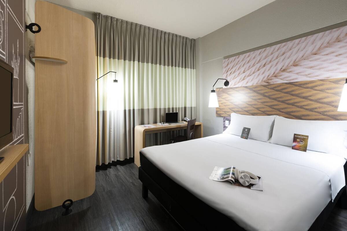 A hotel room with a bed, a desk, a chair, a nightstand, a lamp, and a wall-mounted TV.