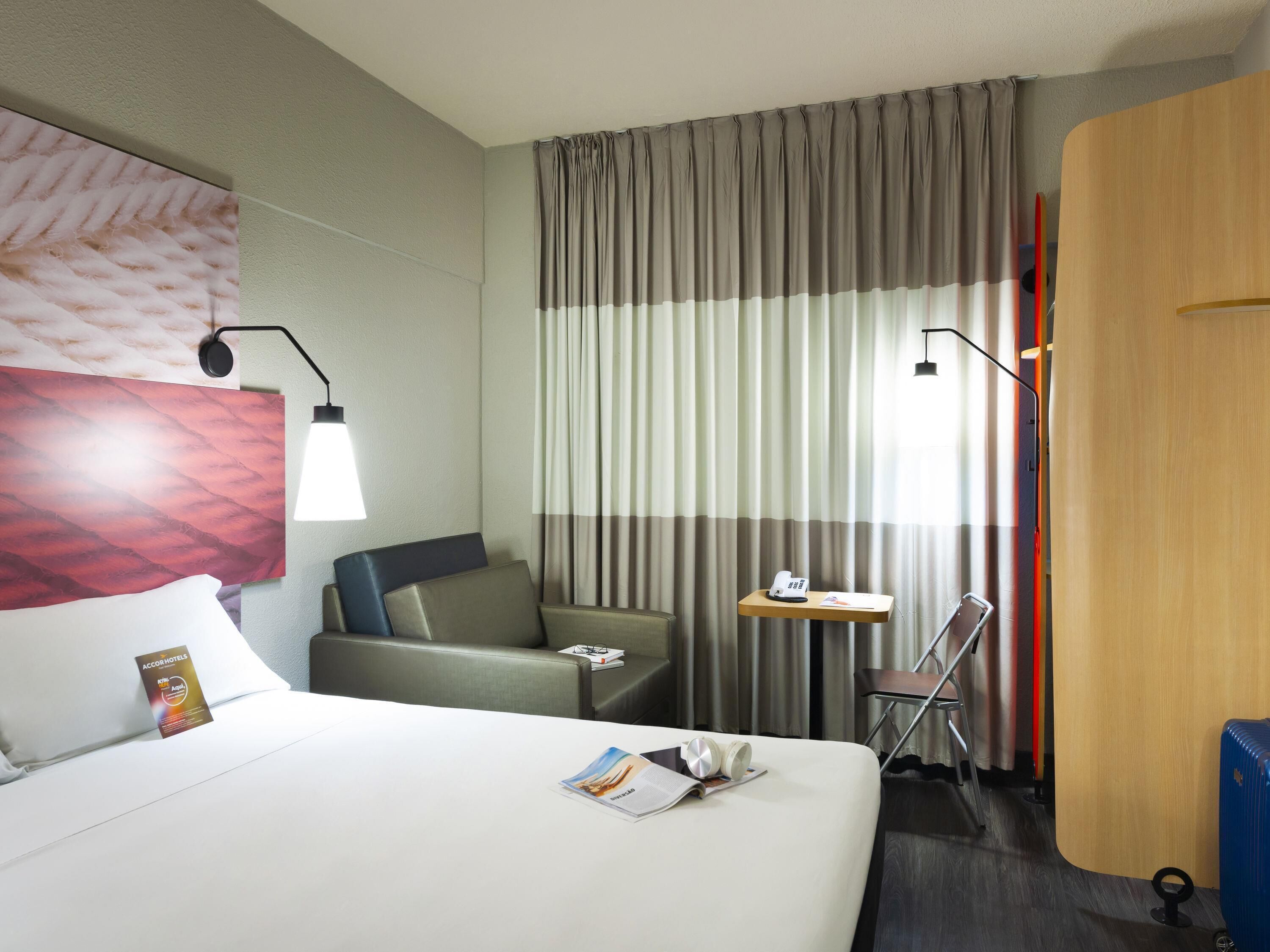 A hotel room with a bed, a desk, a chair, a lamp, and a curtain.