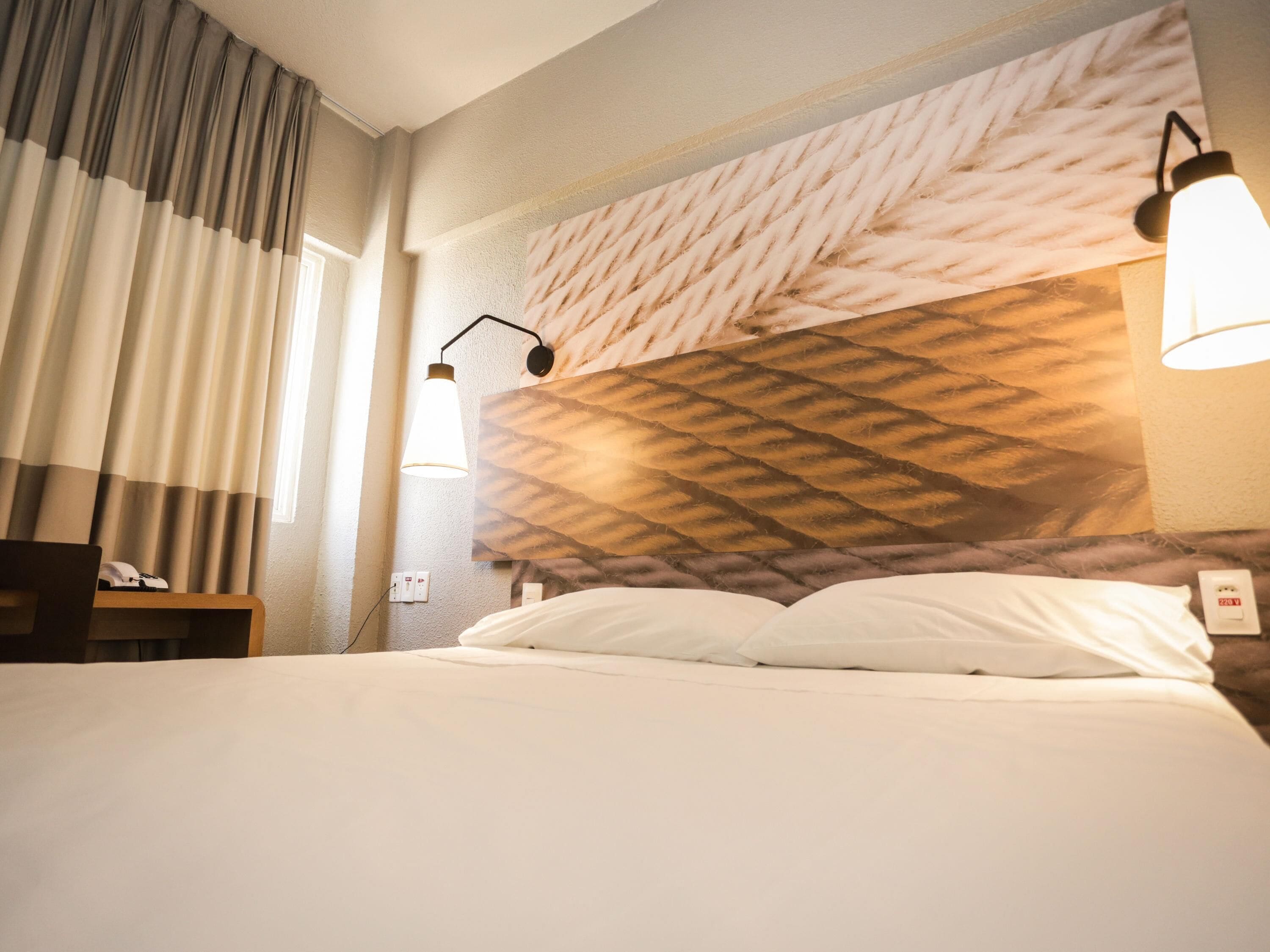 A hotel room with a bed, a headboard with a textured pattern, a nightstand, a lamp, and a window with curtains.