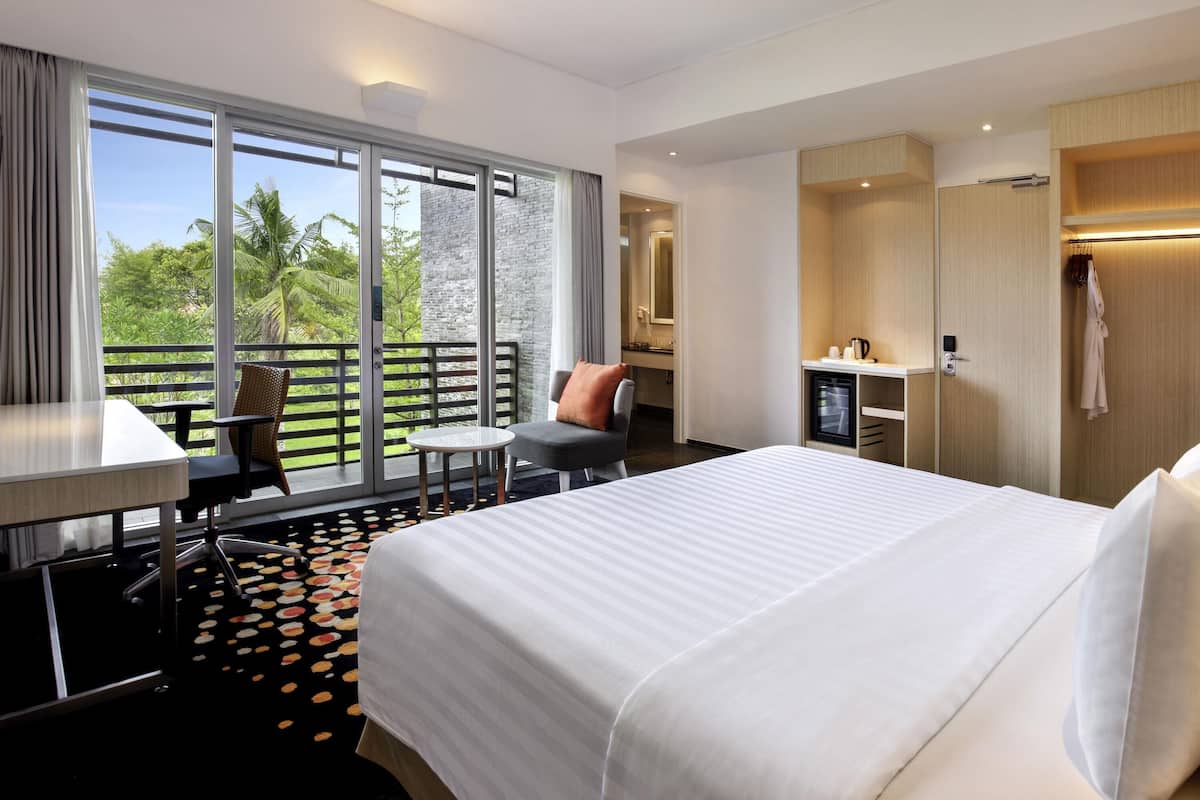 Hotelkamer met een bed, een bureau, een stoel, een klein tafeltje, een minibar en een balkon met uitzicht op groen.