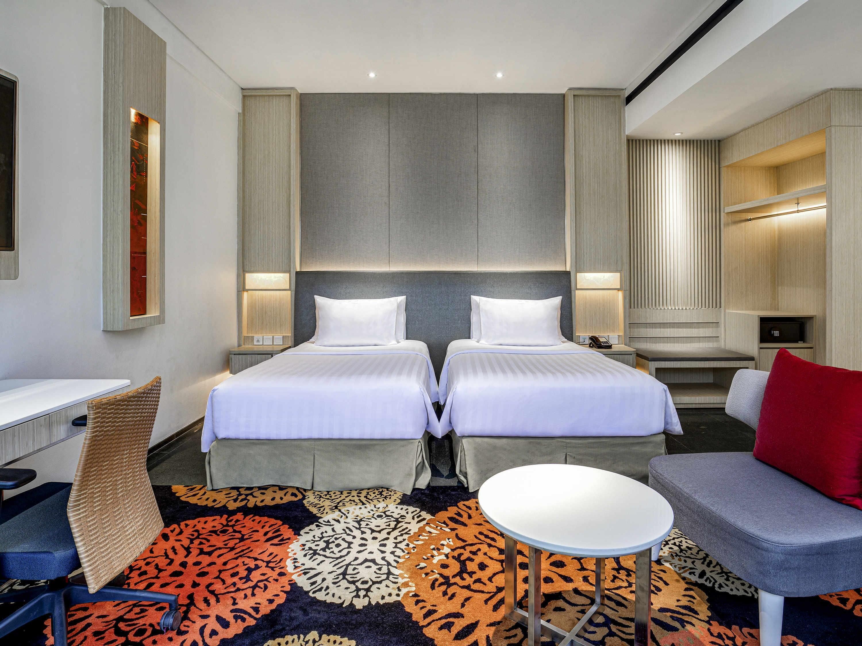 Une chambre d’hôtel avec deux lits, un bureau, une chaise, une petite table et un canapé agrémenté d’un coussin rouge.