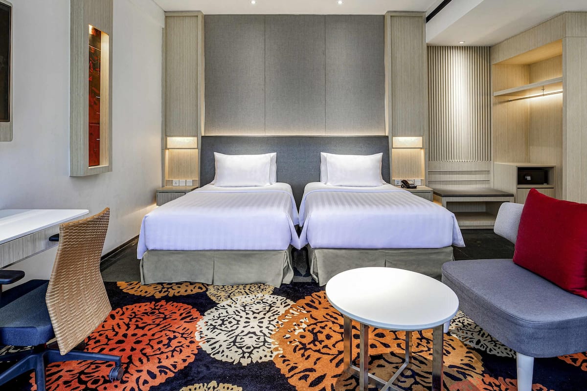 Een hotelkamer met twee bedden, een bureau, een stoel, een kleine tafel en een bank met een rood kussen.
