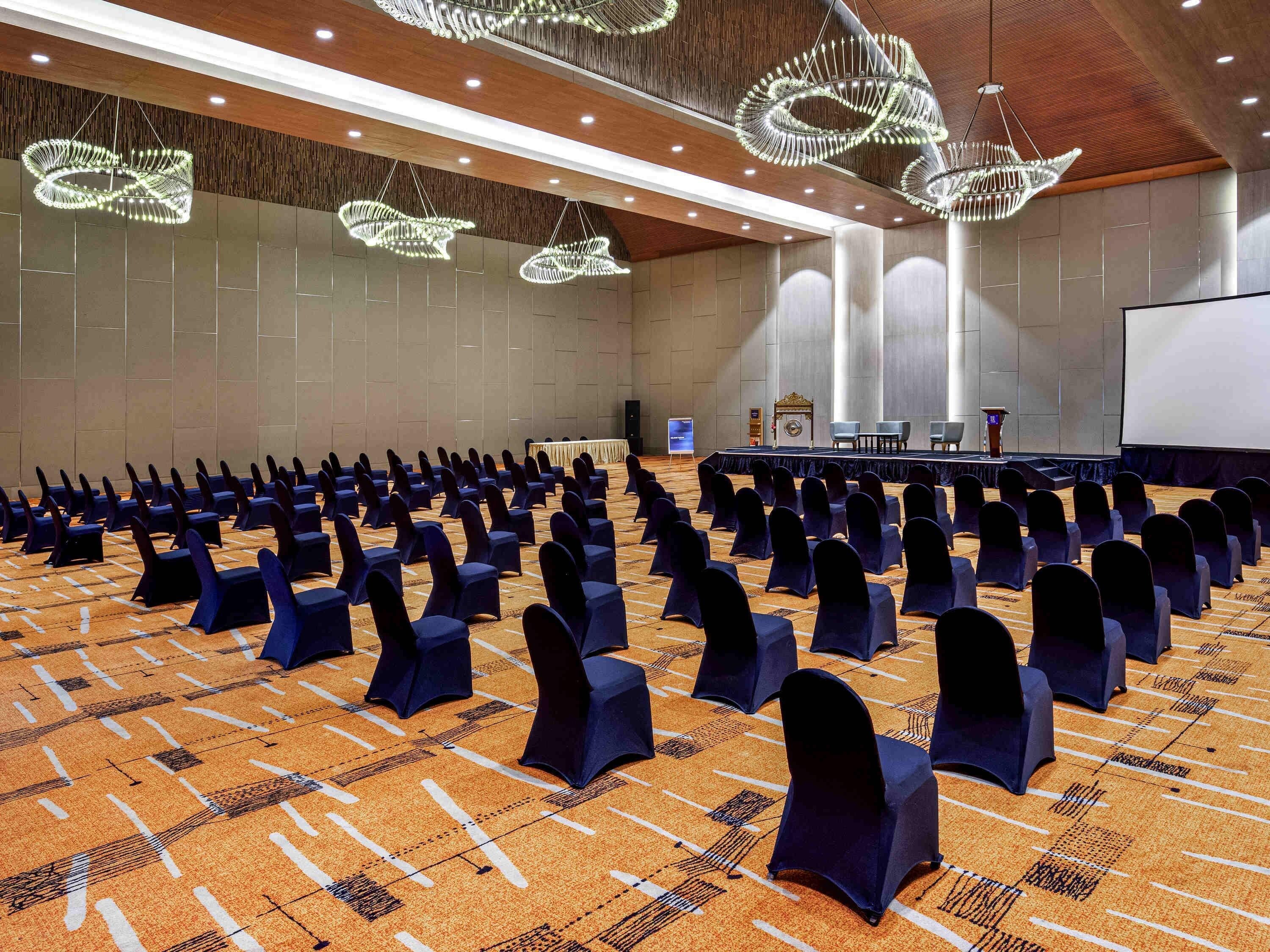 A conference room with rows of chairs, a stage with a podium, and modern chandeliers.