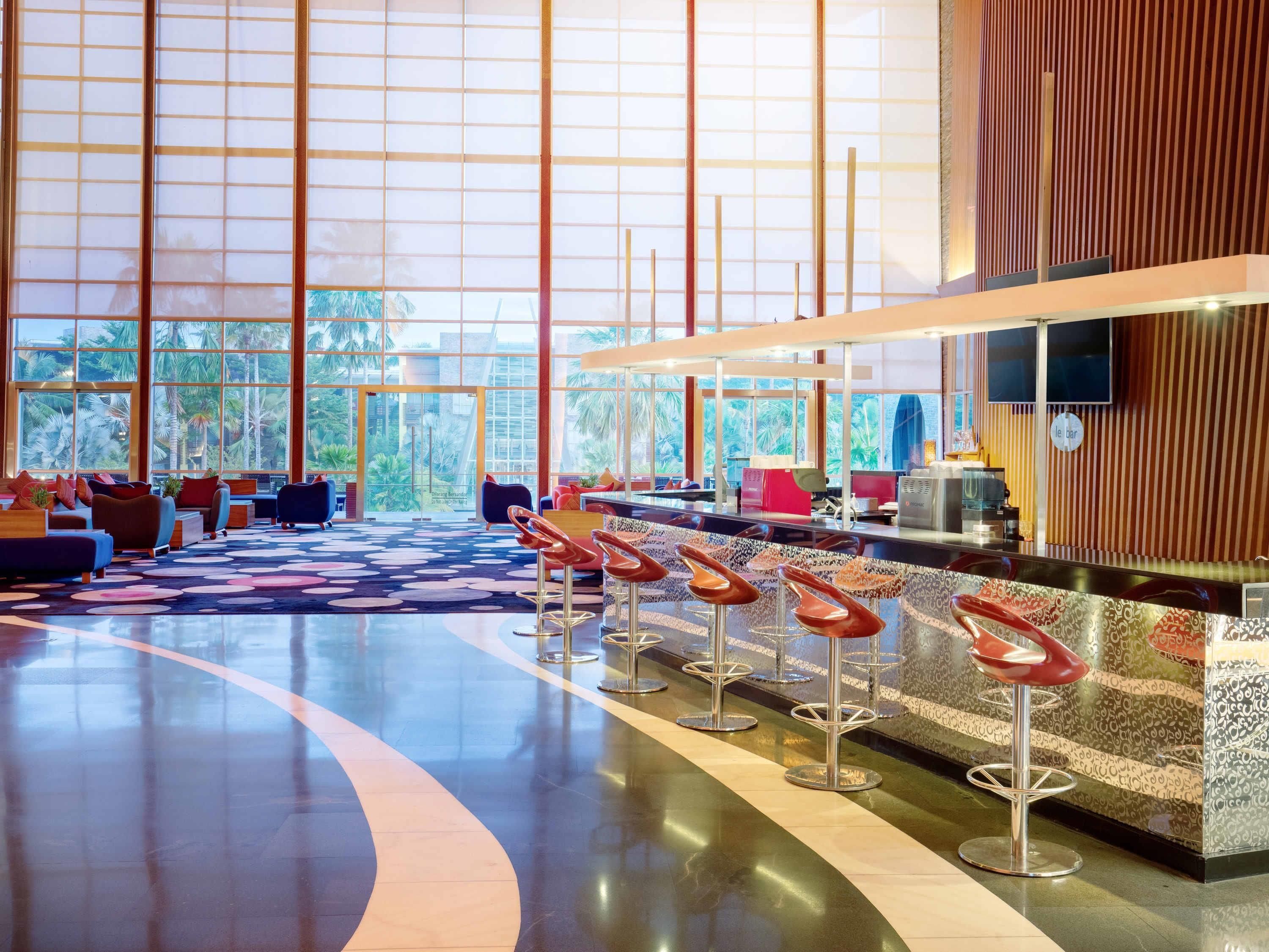 A spacious lobby with a curved seating area, a bar with stools, and large windows offering a view of palm trees and buildings.