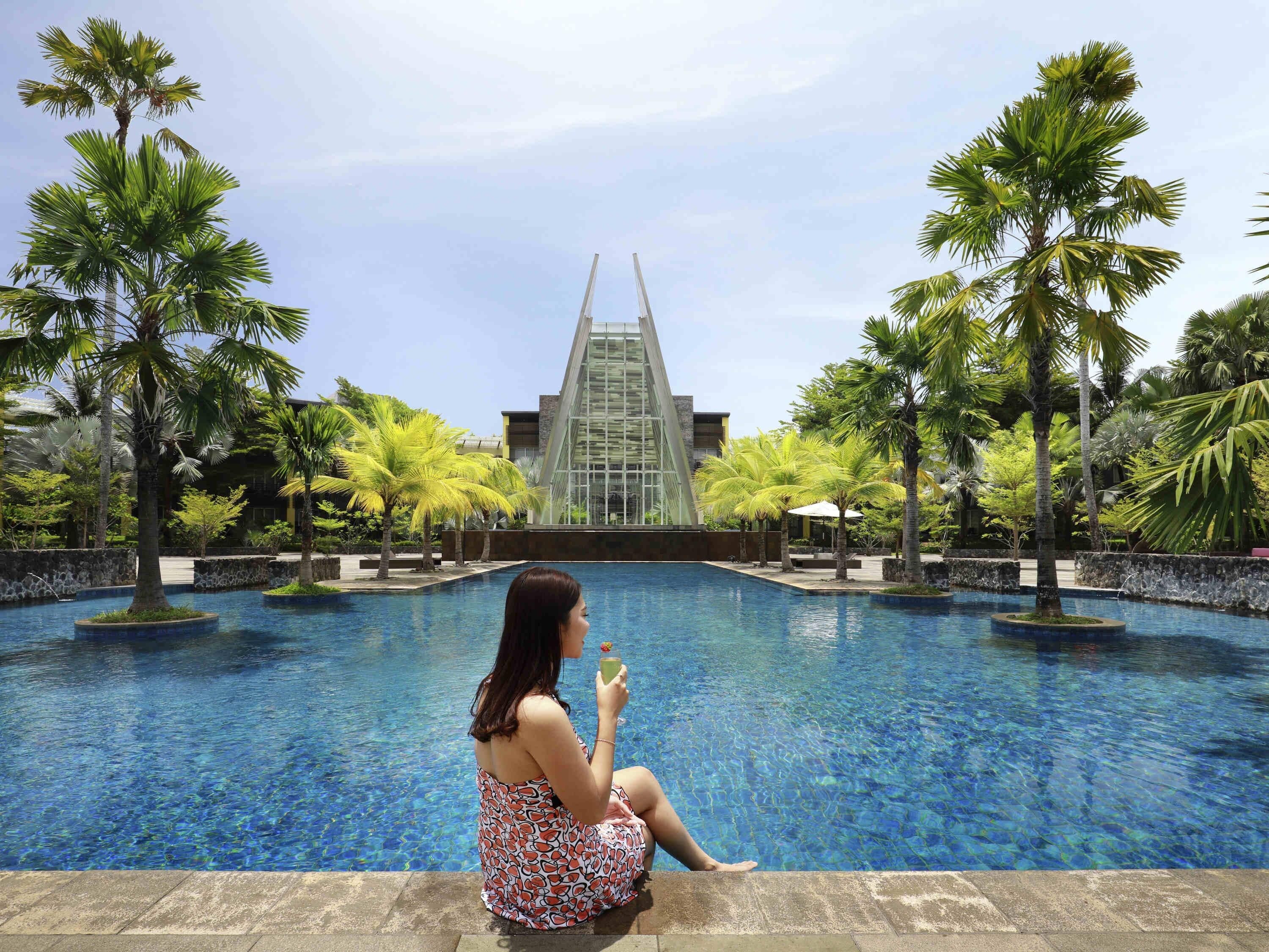 A pool with a woman sitting on the edge, palm trees, and a modern building in the background.