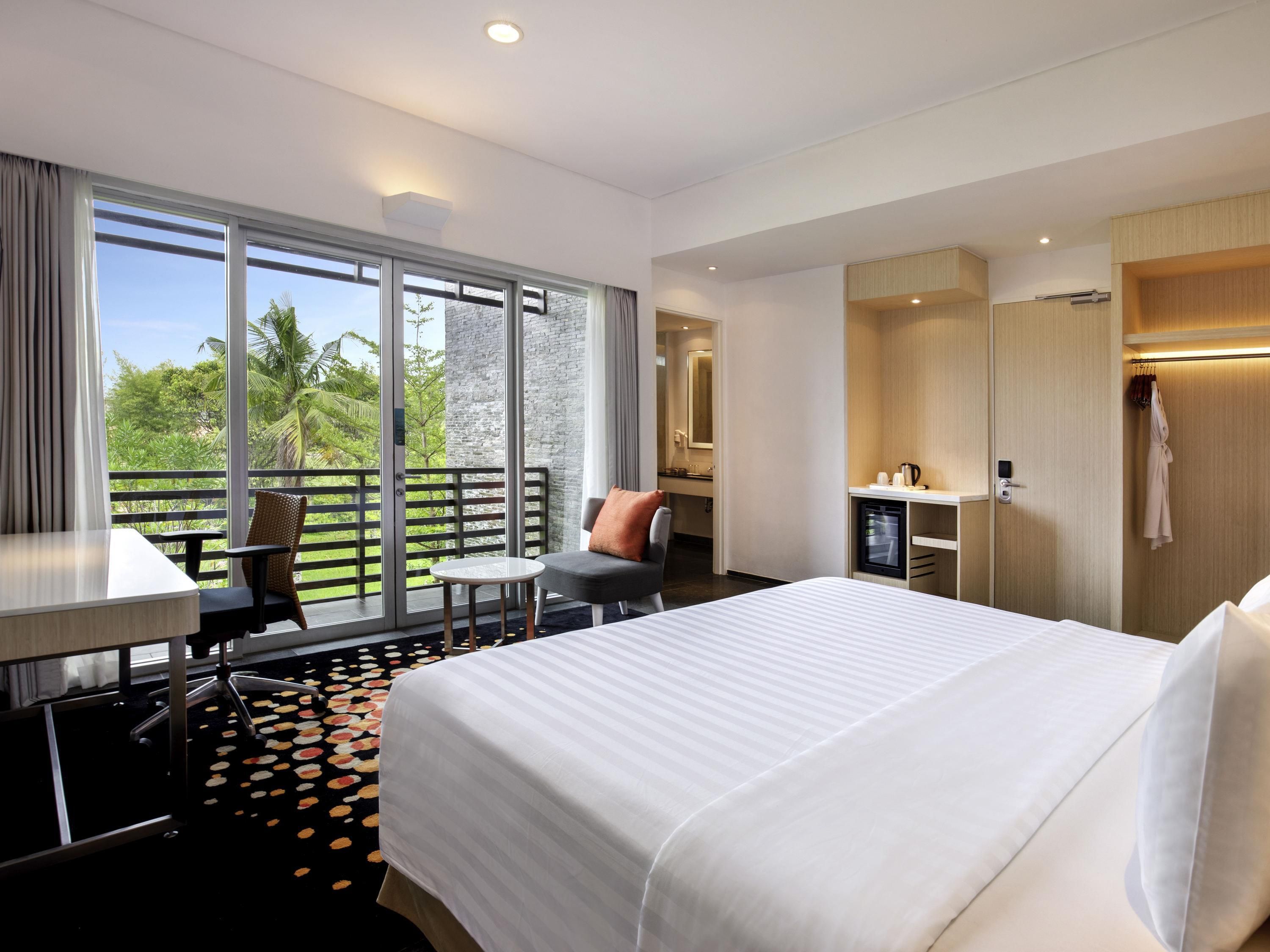 Habitación de hotel con cama, escritorio, silla, una mesita, minibar y balcón con vistas a zonas verdes.