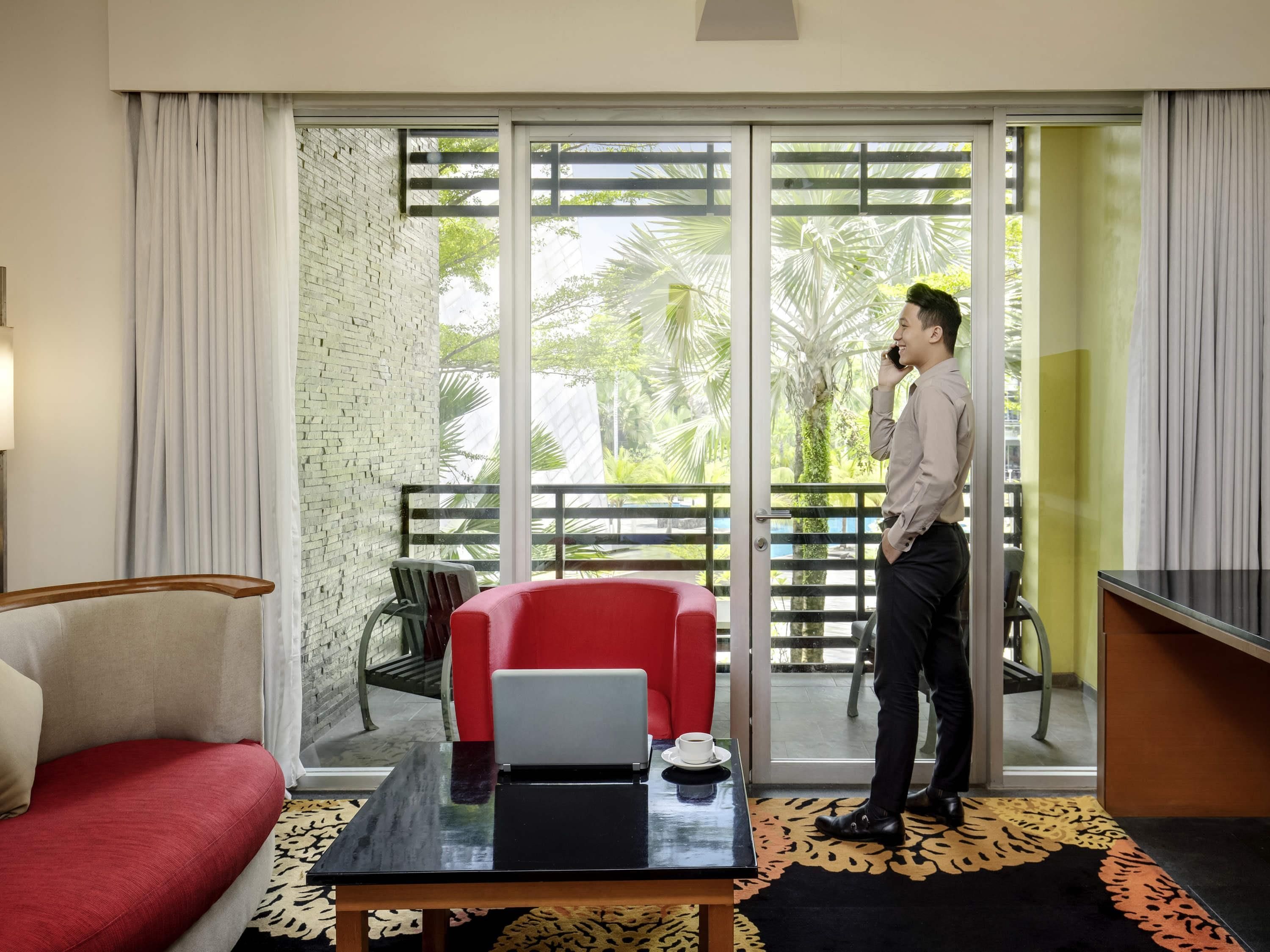 A hotel room with a red sofa, a laptop on a table, and a balcony with a view of greenery.