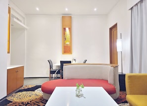 A hotel room with a sofa, a small table with a vase, a desk with chairs, and a large window with curtains.