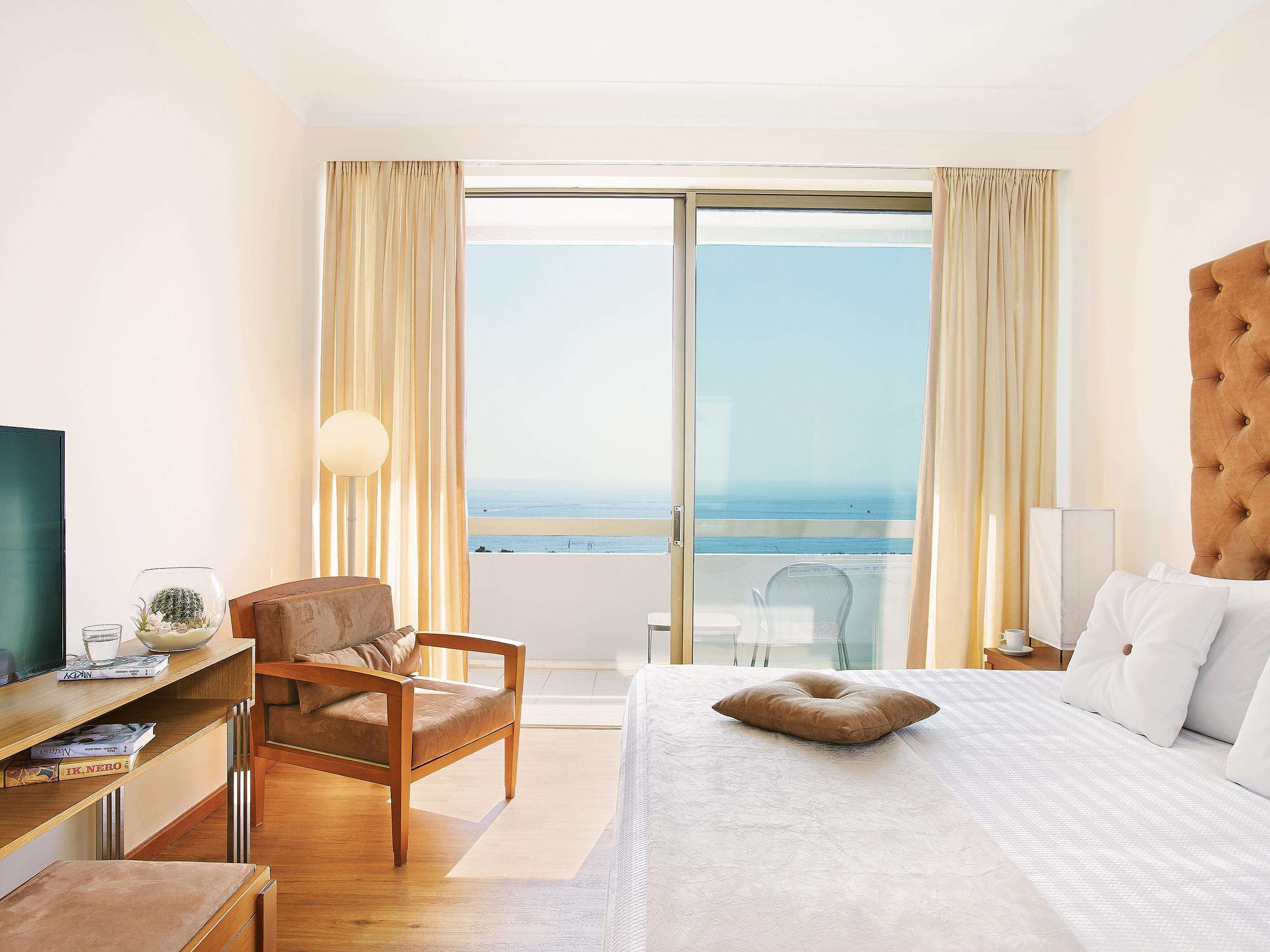 Habitación de hotel con cama, una silla, un escritorio con lámpara y vistas al mar a través de amplias ventanas.