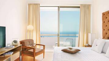 Habitación de hotel con cama, una silla, un escritorio con lámpara y vistas al mar a través de amplias ventanas.