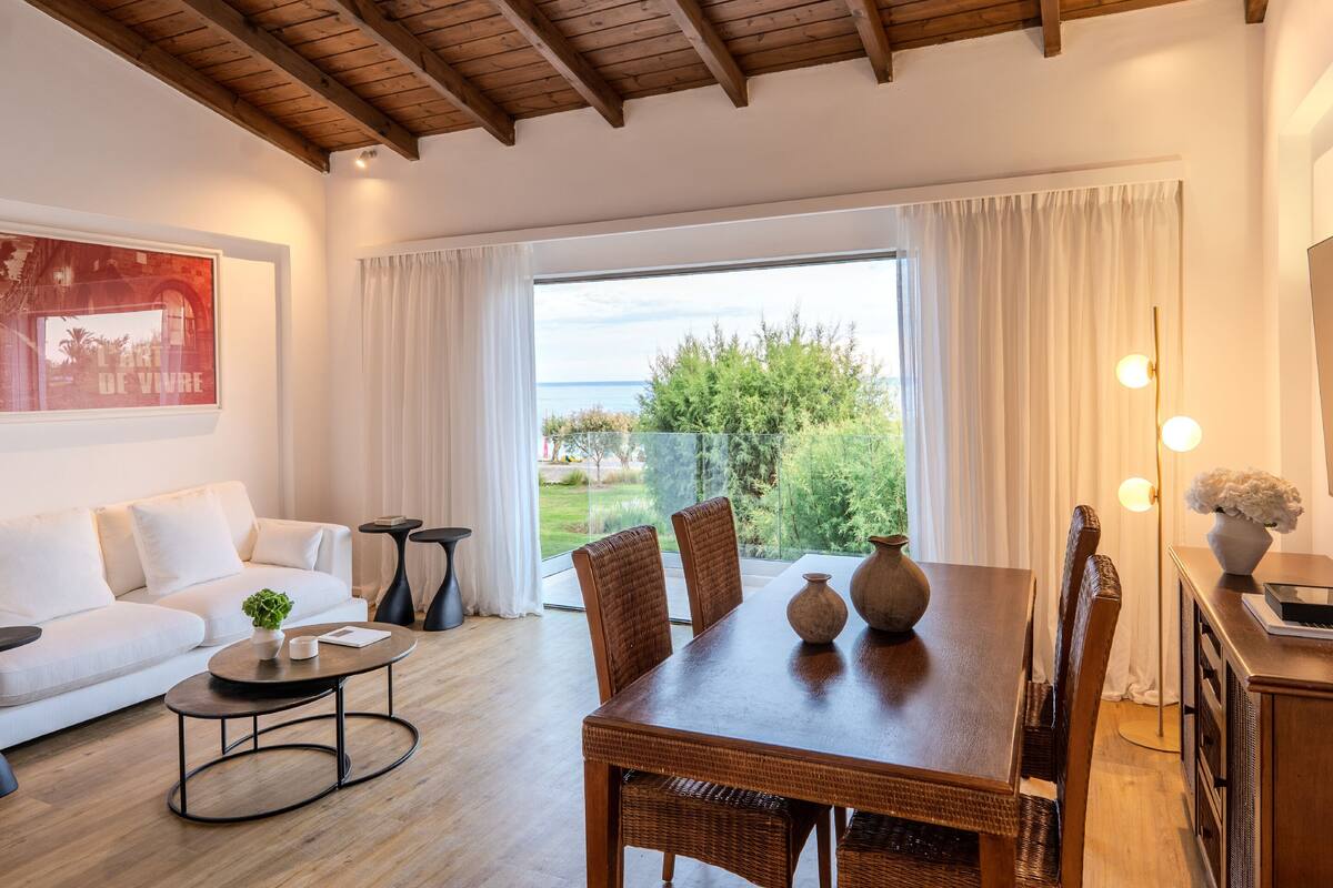 Ein modernes Wohnzimmer mit Holzboden, einem weißen Sofa, einem Esstisch mit Stühlen und Meerblick durch große Fenster.