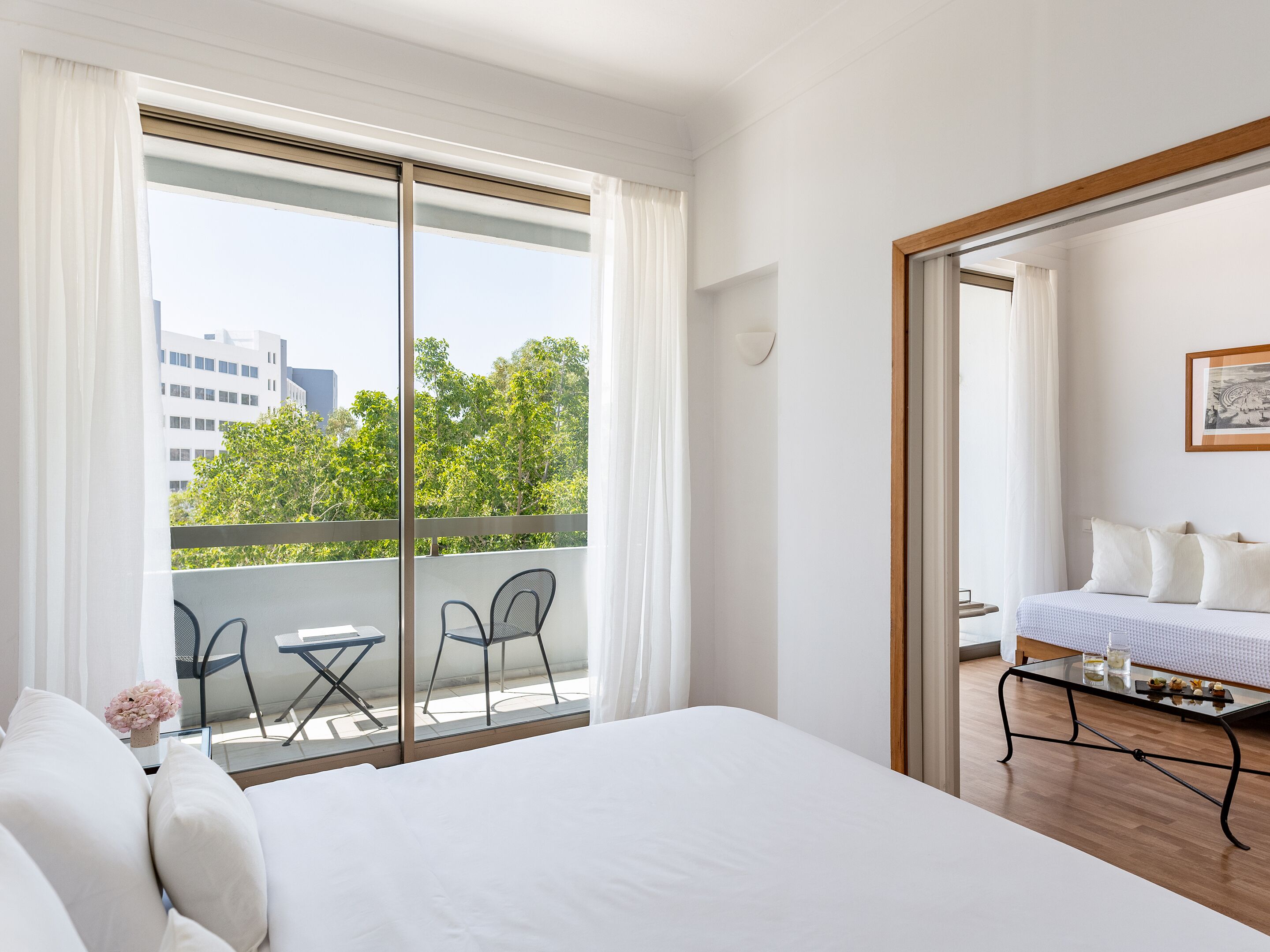 Une chambre d’hôtel avec un grand lit, un balcon doté d’une table et de chaises, et une vue sur les bâtiments et les arbres.