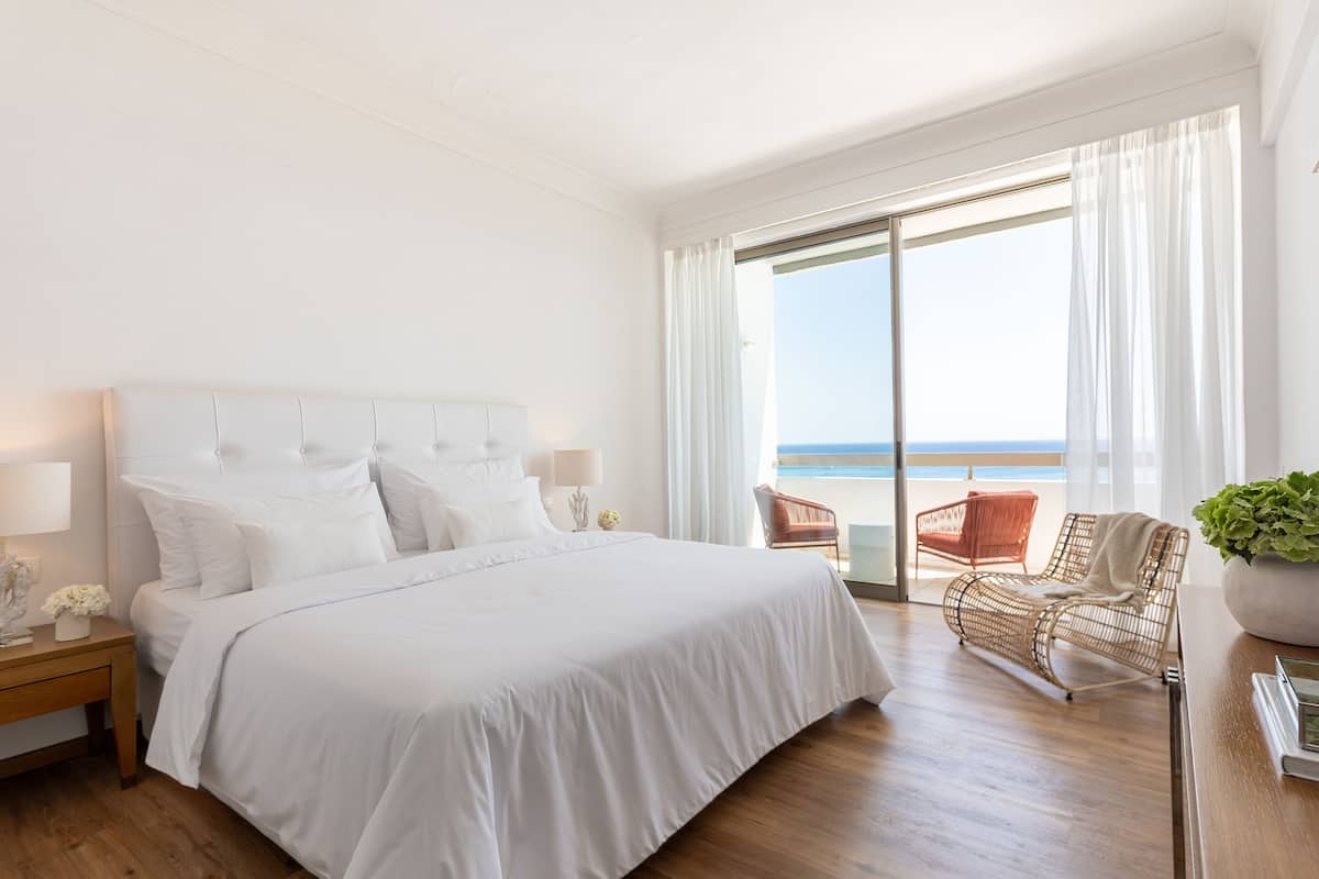 Ein Schlafzimmer mit einem großen Bett, einem Nachttisch, einer Lampe und Blick auf den Strand durch ein großes Fenster.