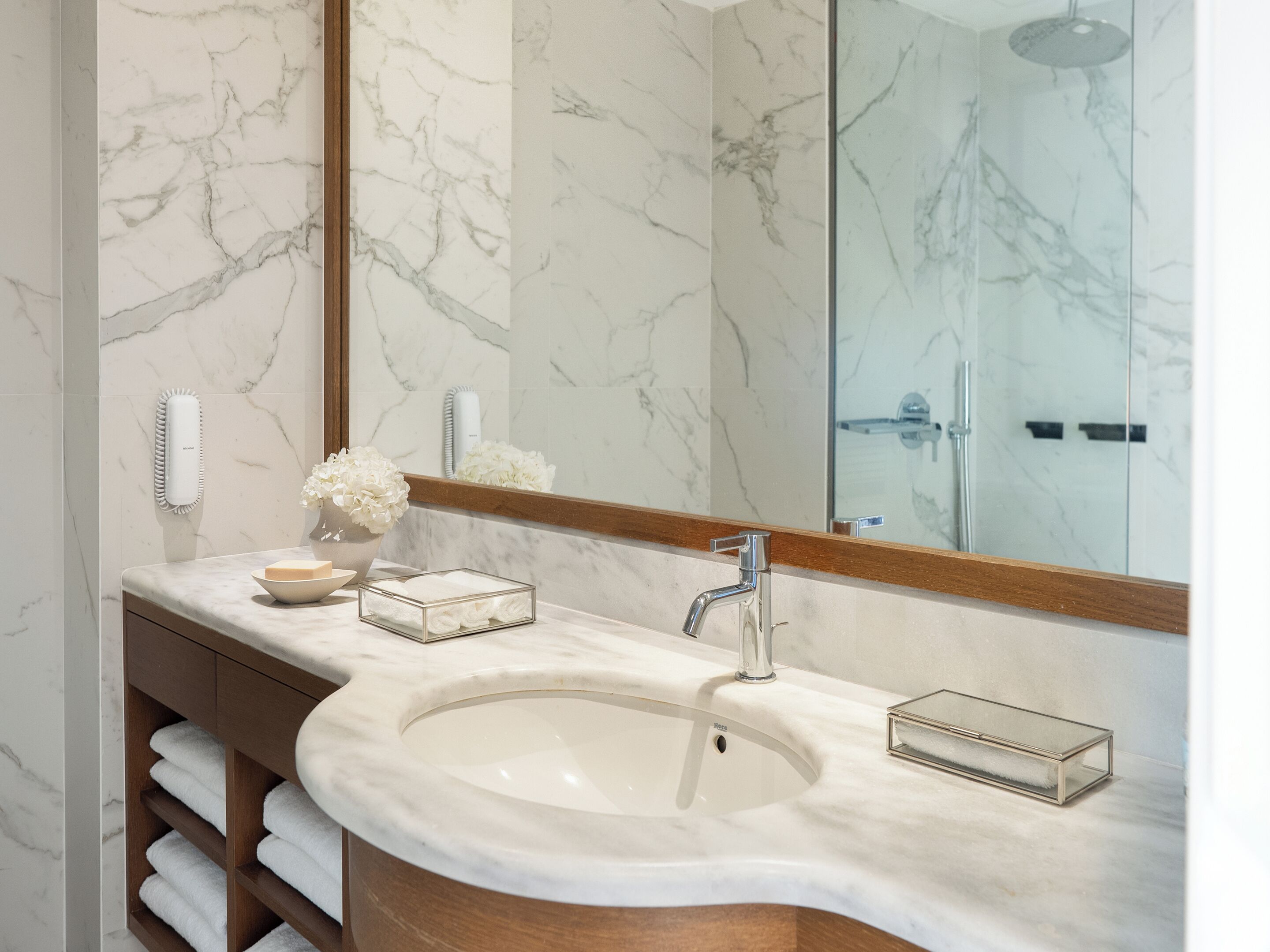 Un baño con lavamanos de mármol, tocador de madera, espejo y cabina de ducha de vidrio.