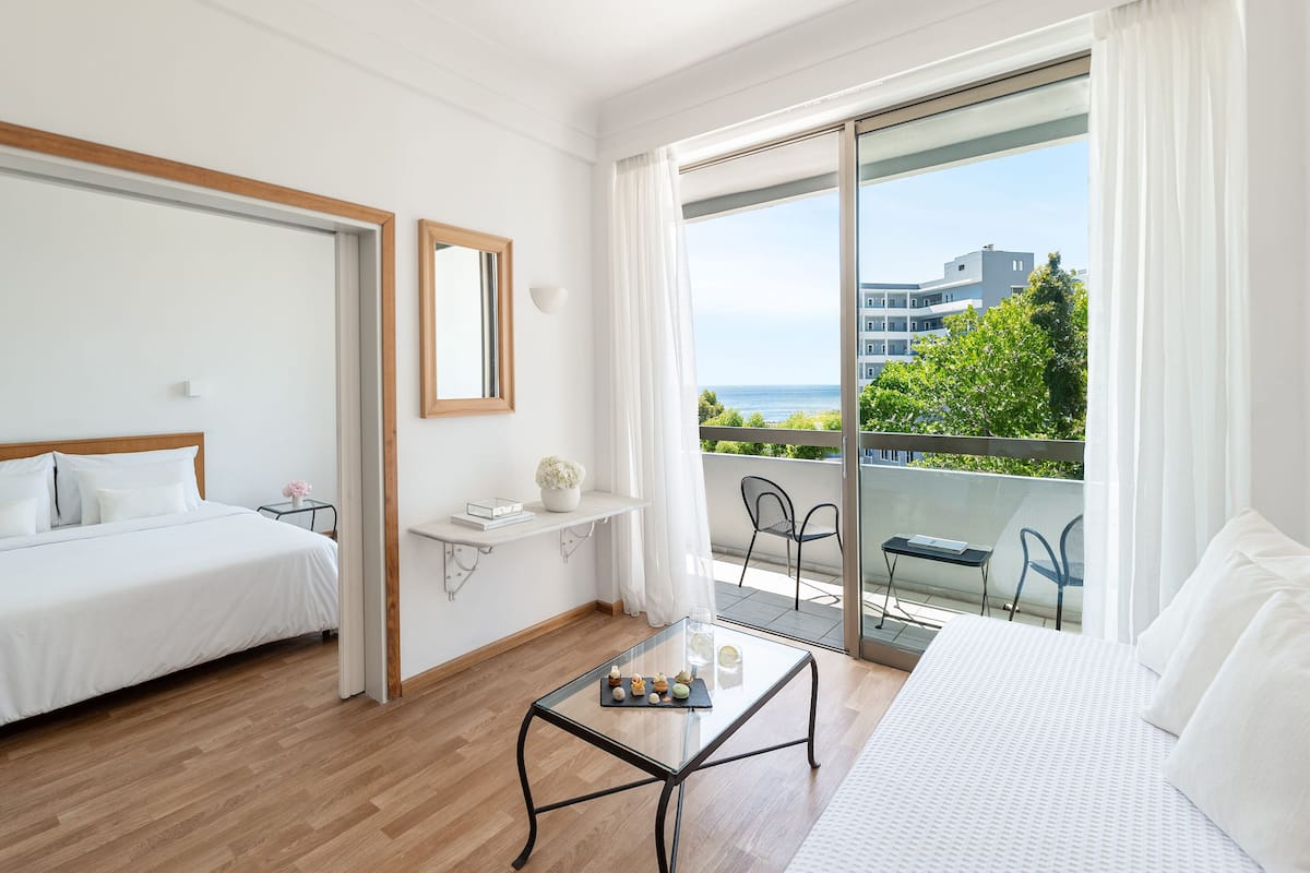 Ein Hotelzimmer mit Bett, einem kleinen Tisch mit Snacks, einem Sessel, einer Couch und einem Balkon mit Blick auf Gebäude und Grünflächen.