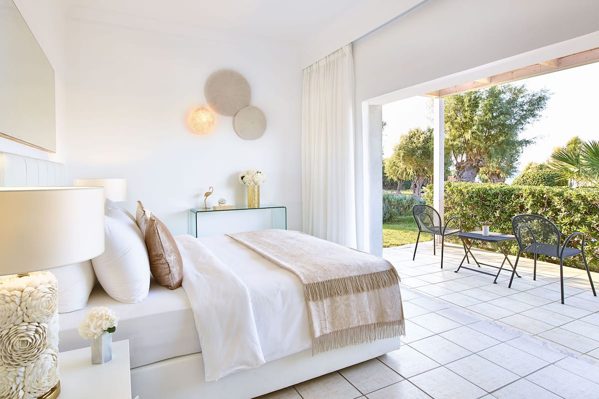 Ein modernes Schlafzimmer mit einem großen Bett, Nachttischen, einem Beistelltisch und Blick auf eine Terrasse mit Sitzgelegenheiten und viel Grün.