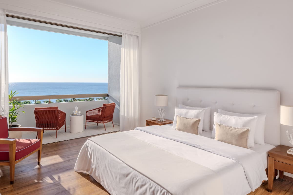 Ein Hotelzimmer mit einem großen Bett, einem Holfnachtschränkchen, einem Sessel, einem kleinen Tisch und Blick aufs Meer.