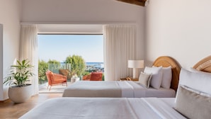 Habitación de hotel con dos camas, un ventanal con cortinas, una planta en maceta, sillas de mimbre y vista al mar.