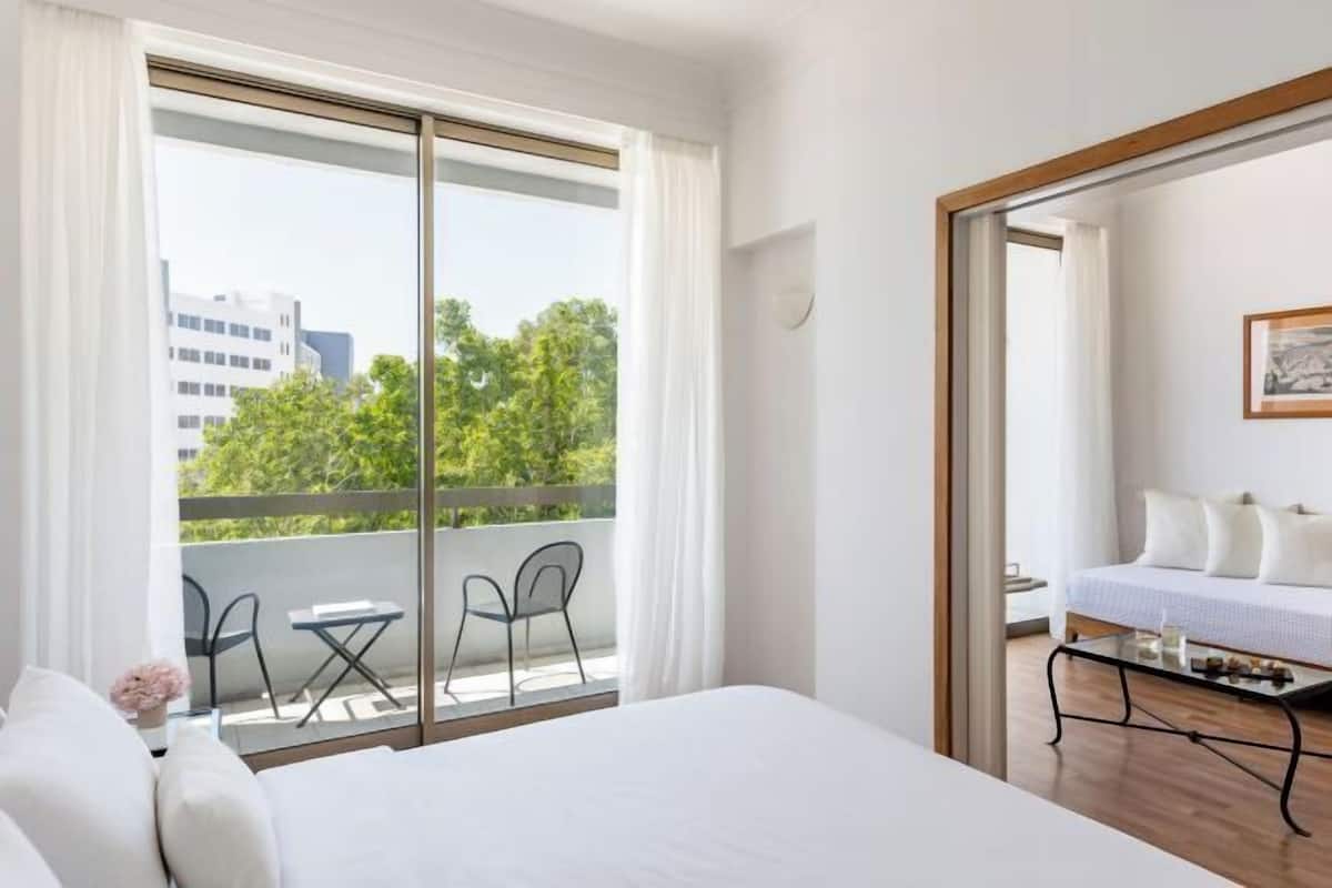 Ein Hotelzimmer mit einem großen Bett, einem Balkon mit Stühlen und Blick auf Gebäude und Bäume.