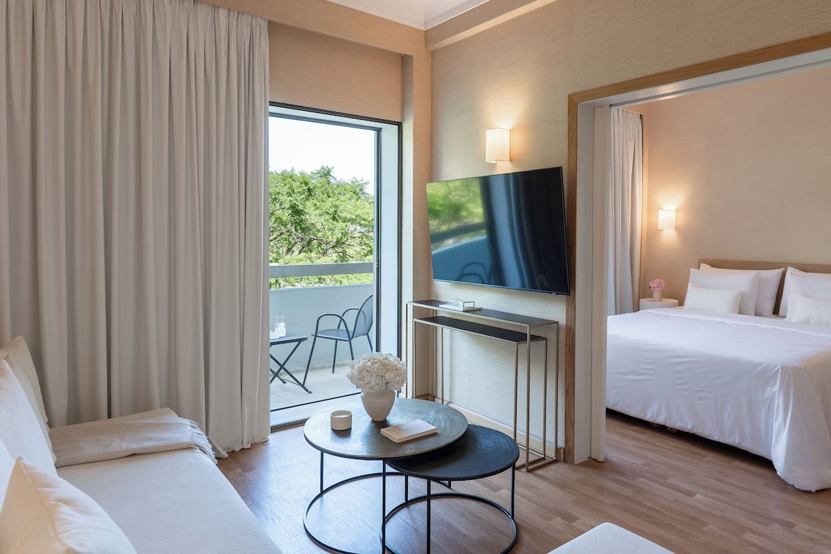 Ein Hotelzimmer mit Bett, Fernseher, einem kleinen Tisch mit einer Vase und einem Balkon mit Blick auf Bäume.