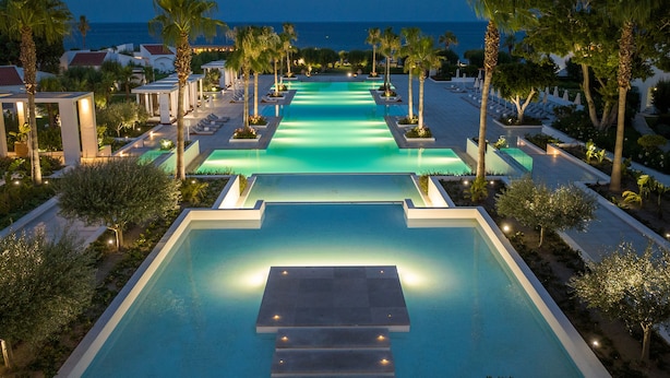 Zona de piscina con varias piscinas, palmeras y vista al océano al atardecer.