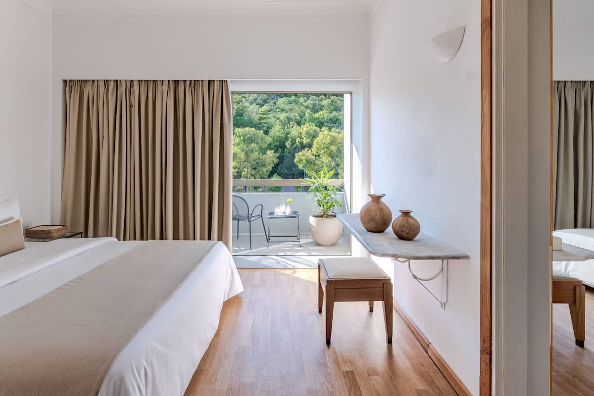 Ein Hotelzimmer mit einem großen Bett, einem kleinen Tisch mit zwei Vasen, einer Topfpflanze und Blick auf Grünfläche draußen.