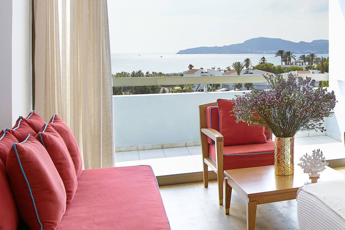 Ein Balkon mit einer roten Couch und Stühlen, einem Holztisch mit Blumen und Blick auf das Meer und die entfernten Hügel.