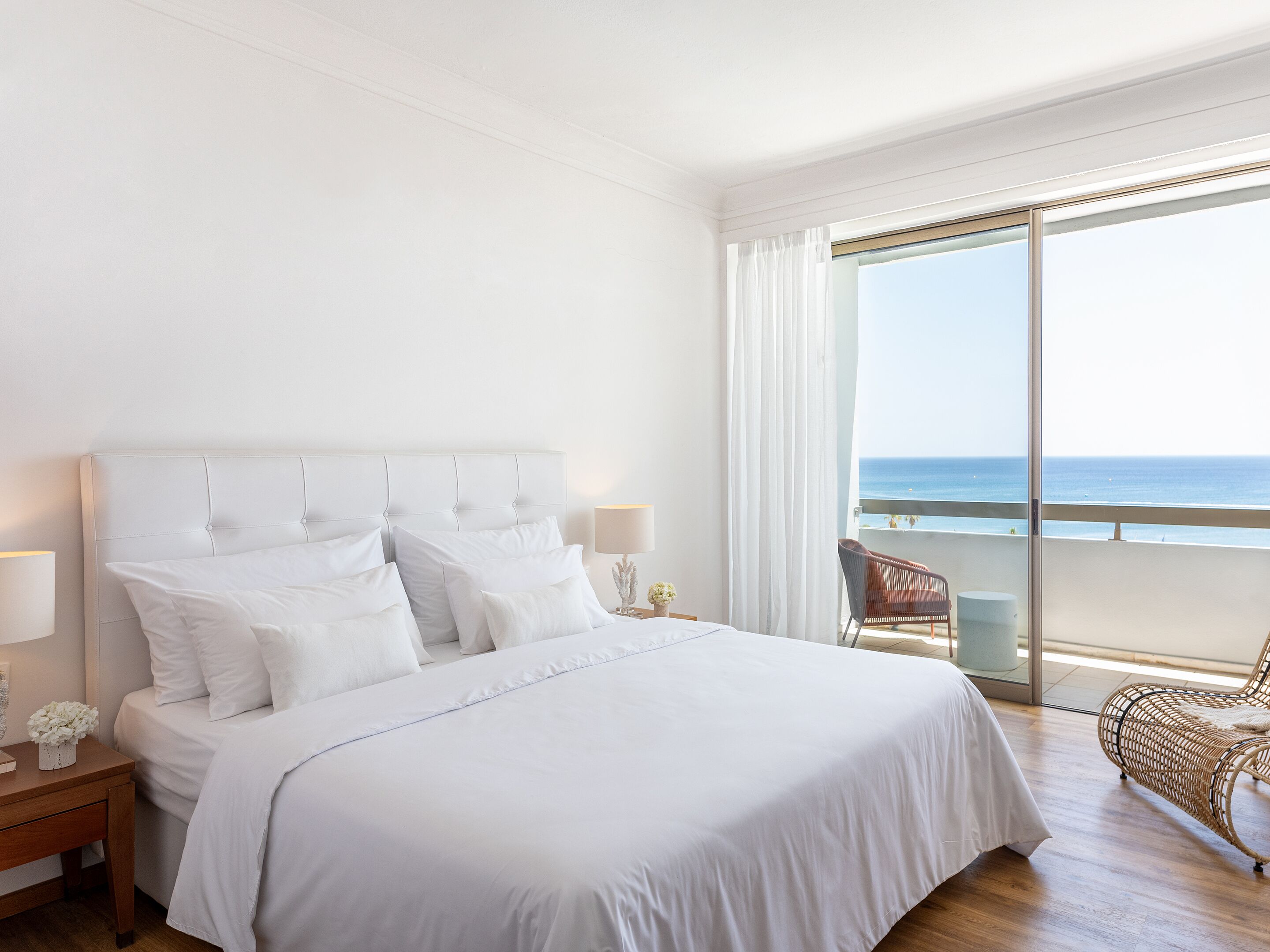 Une chambre d’hôtel avec un grand lit, une table de chevet, une lampe, un fauteuil en osier et un balcon donnant sur la mer.