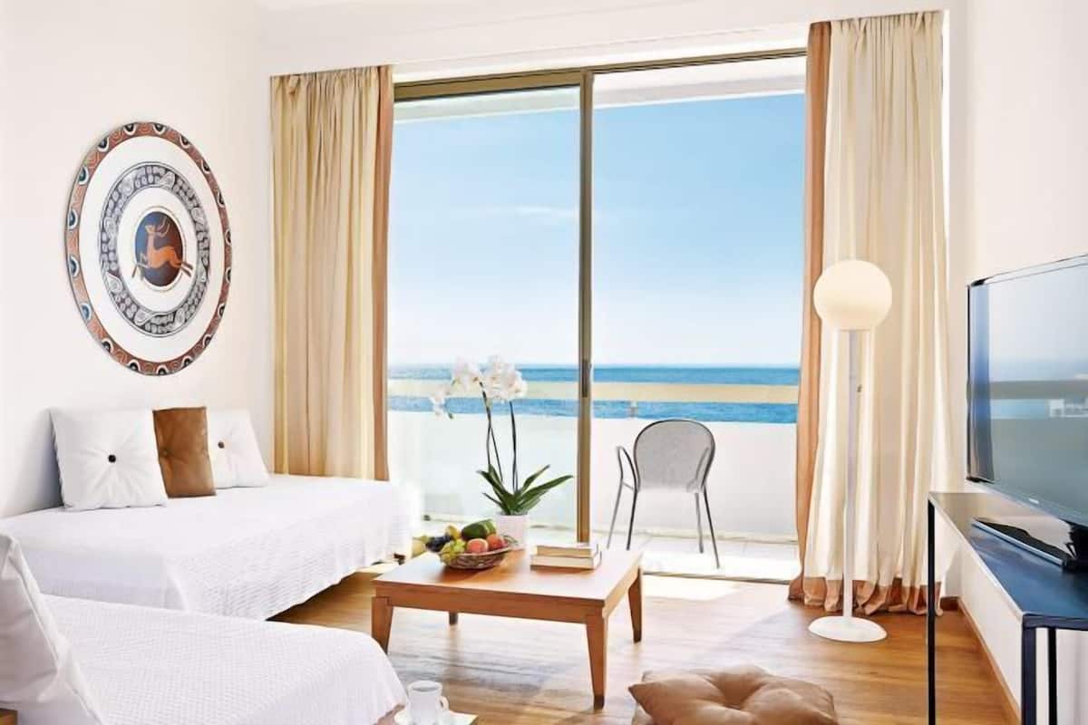 Ein Hotelzimmer mit Bett, Fernseher, Tisch mit Obstschale, Stuhl und Blick auf den Ozean.