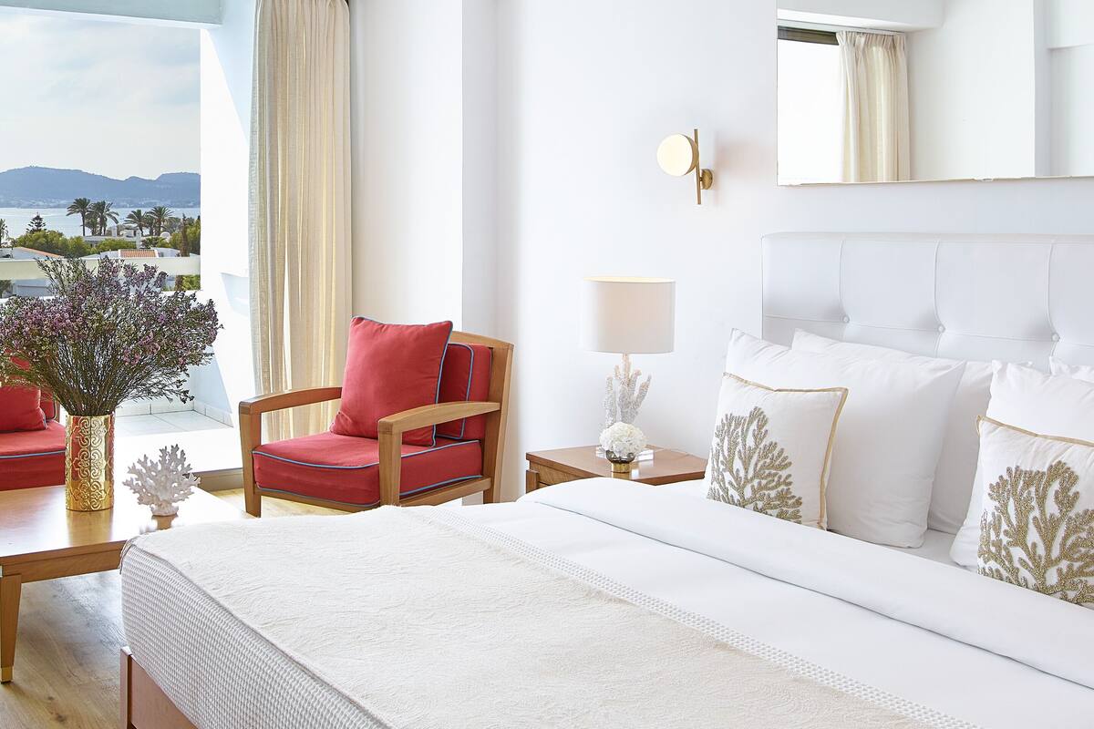 Ein Hotelzimmer mit Bett, Sessel, Nachttisch, Lampe und Meerblick.