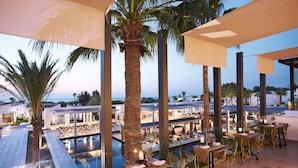 Un área de comedor en la azotea con mesas y sillas, con vista a una piscina y al mar.