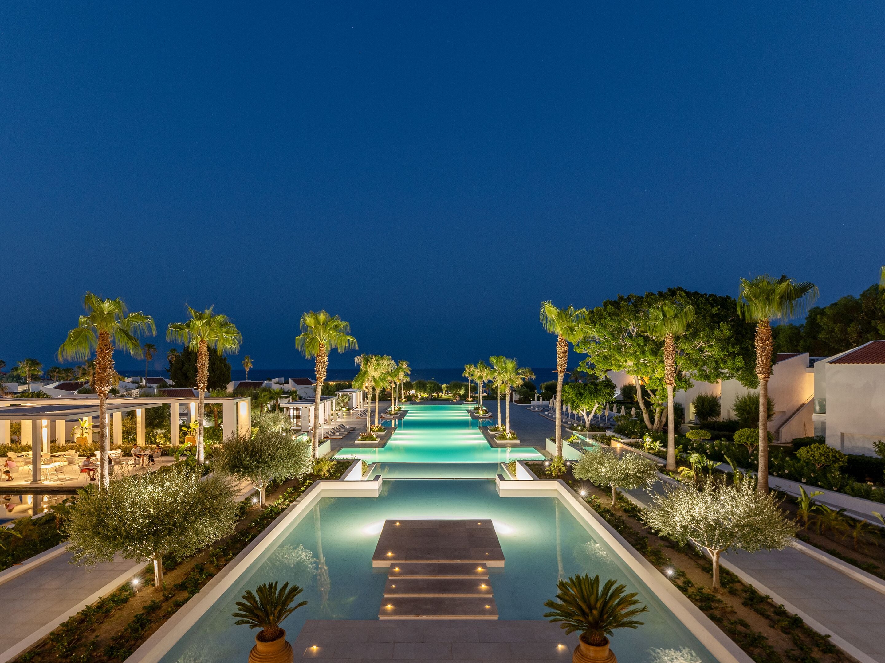 A nighttime resort pool area with a clear view of the ocean, surrounded by palm trees and modern buildings.