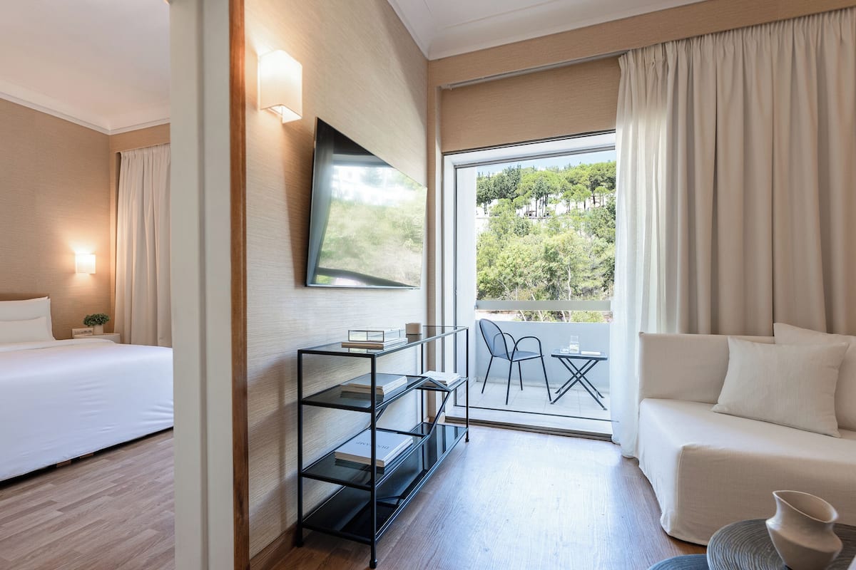 Ein modernes Hotelzimmer mit Bett, Sofa, Fernseher, Glastisch und einem Balkon mit Blick auf Bäume.