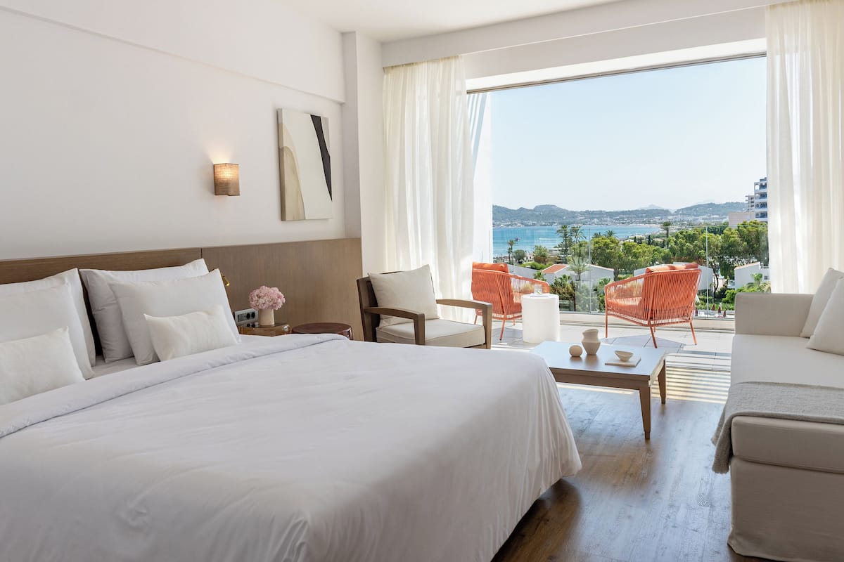 Ein Hotelzimmer mit einem großen Bett, einer Sitzecke mit zwei Sesseln, einem kleinen Tisch und Blick auf das Meer und die Hügel.