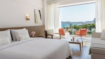 A hotel room with a large bed, a sitting area with two chairs, a small table, and a view of the sea and hills.