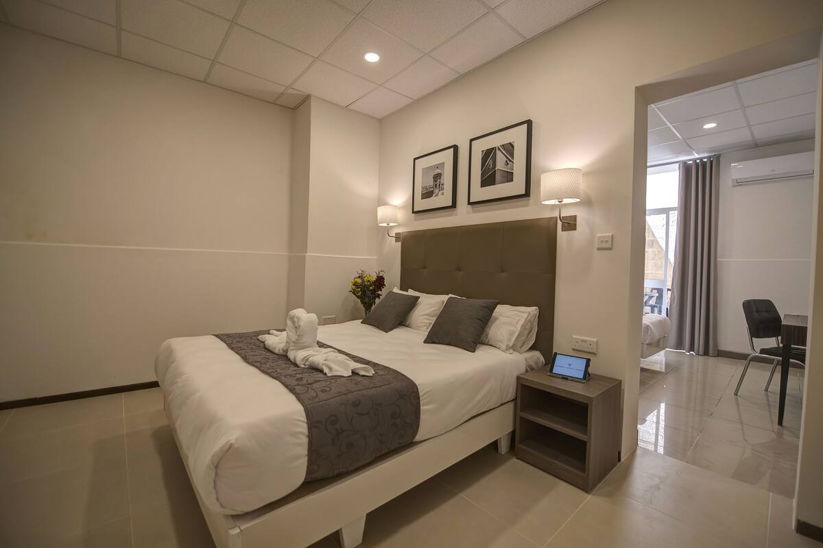 Ein modernes Hotelzimmer mit einem großen Bett, einem Nachttisch mit einem Laptop und Blick auf das angrenzende Zimmer.