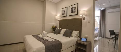 A modern hotel room with a large bed, a bedside table with a laptop, and a view of the adjacent room.