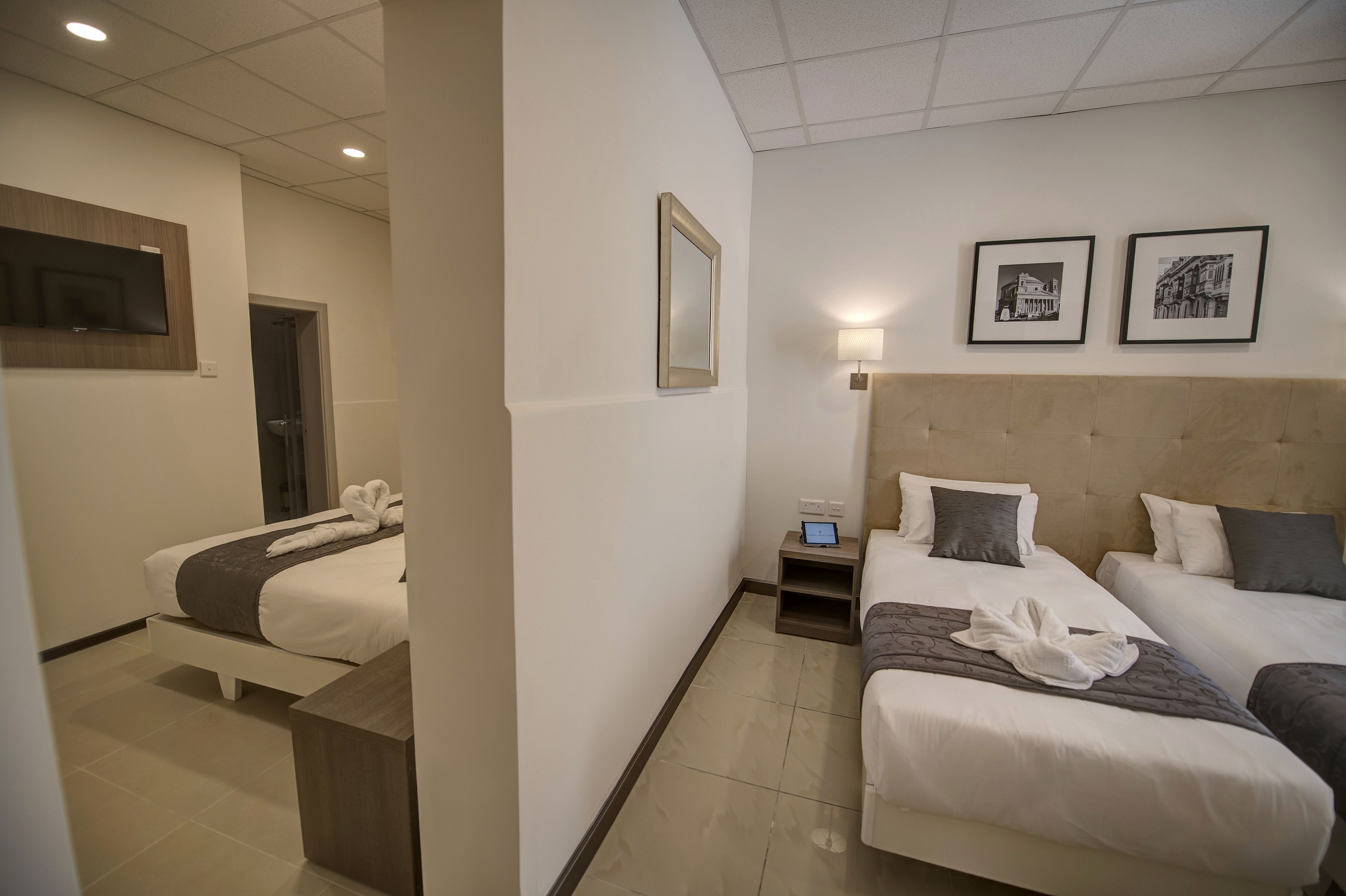 Een hotelkamer met twee eenpersoonsbedden, een nachtkastje met een lamp, een tv aan de muur en ingelijste foto's.