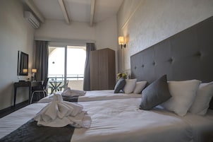 A hotel room with two beds, a desk with a chair, a small table, and a balcony with a view.