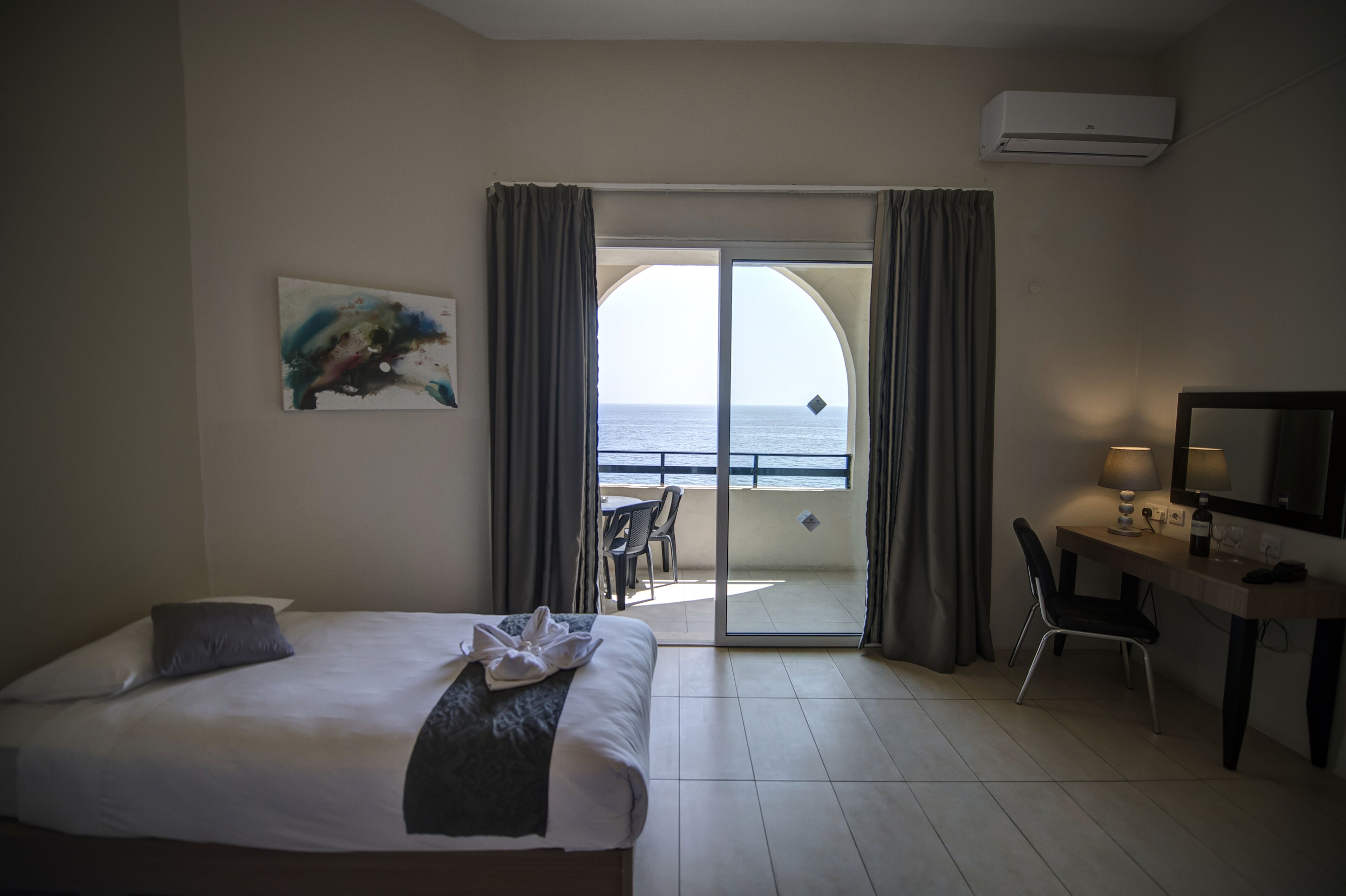 Une chambre d’hôtel avec un lit, un bureau, une chaise, un tableau et une vue sur la mer, visible à travers une porte vitrée.