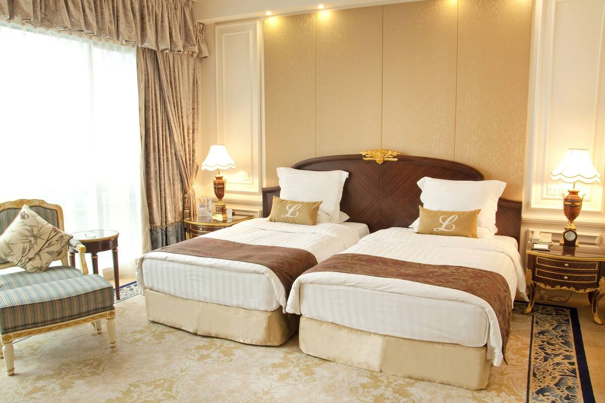 A hotel room with two beds, a chair, and two bedside tables with lamps.