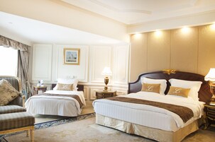 Habitación de hotel con dos camas, una zona de estar con una silla, una mesita de noche con lámpara y un cuadro en la pared.