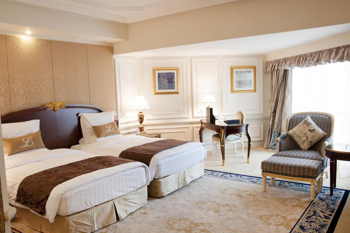 A hotel room with two beds, a desk, a chair, and a large window with curtains.