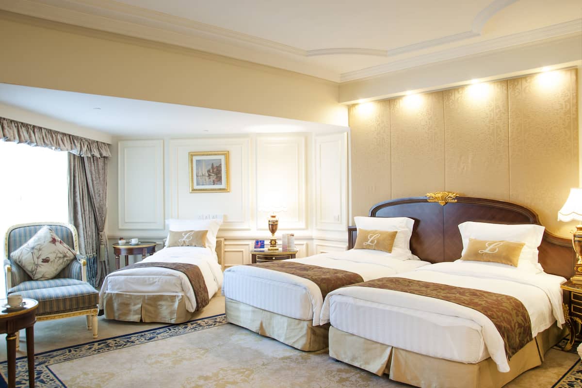 A hotel room with two beds, a sitting area with a chair, a small table, and a nightstand with a lamp.