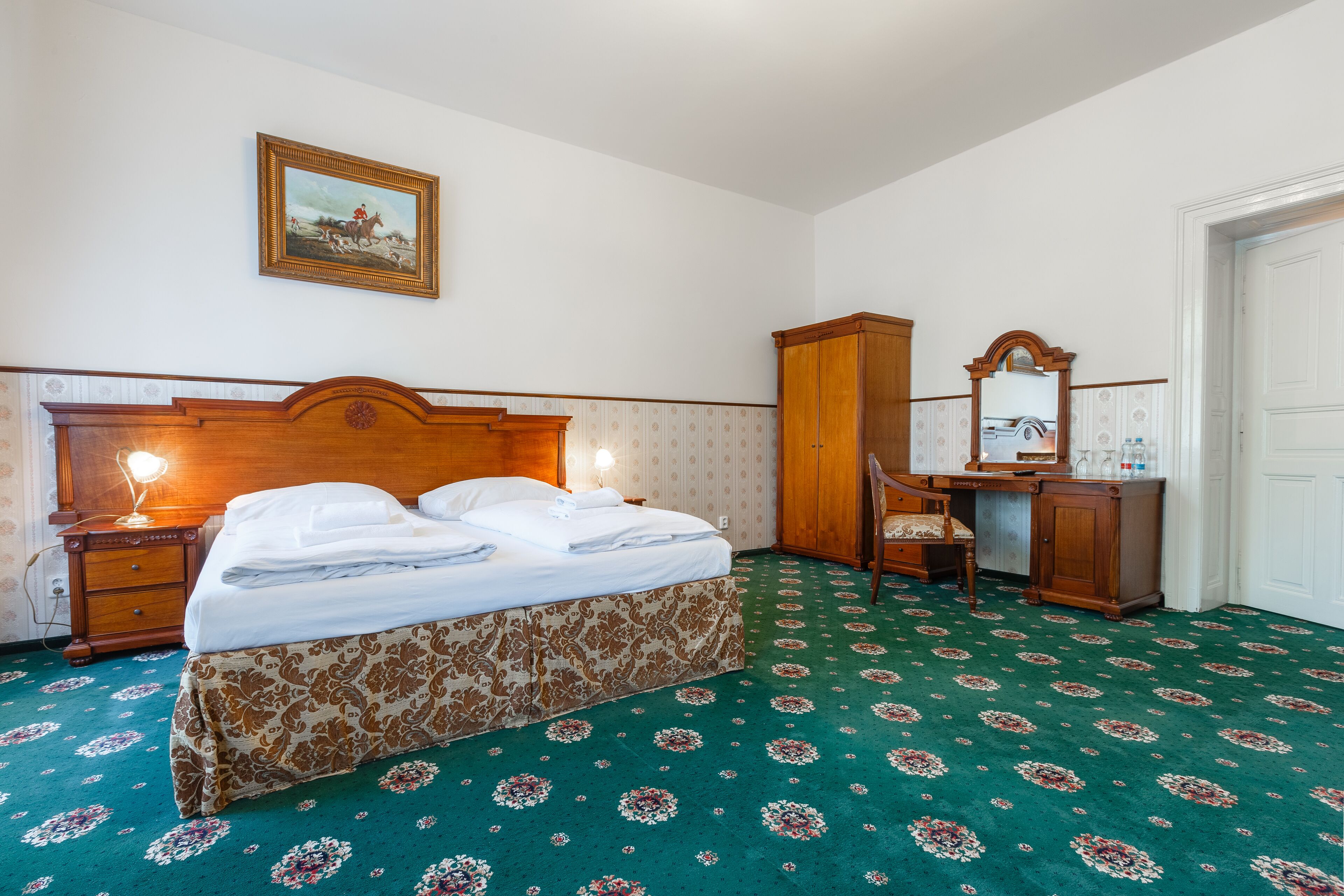A hotel room with a bed, a desk, a chair, a wardrobe, a mirror, and a painting on the wall.