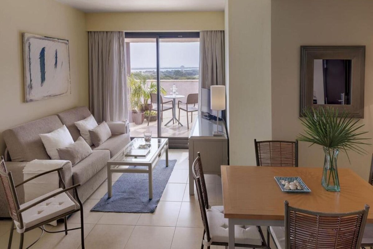 A hotel room with a sofa, coffee table, dining table, chairs, a balcony with a view, and a painting on the wall.