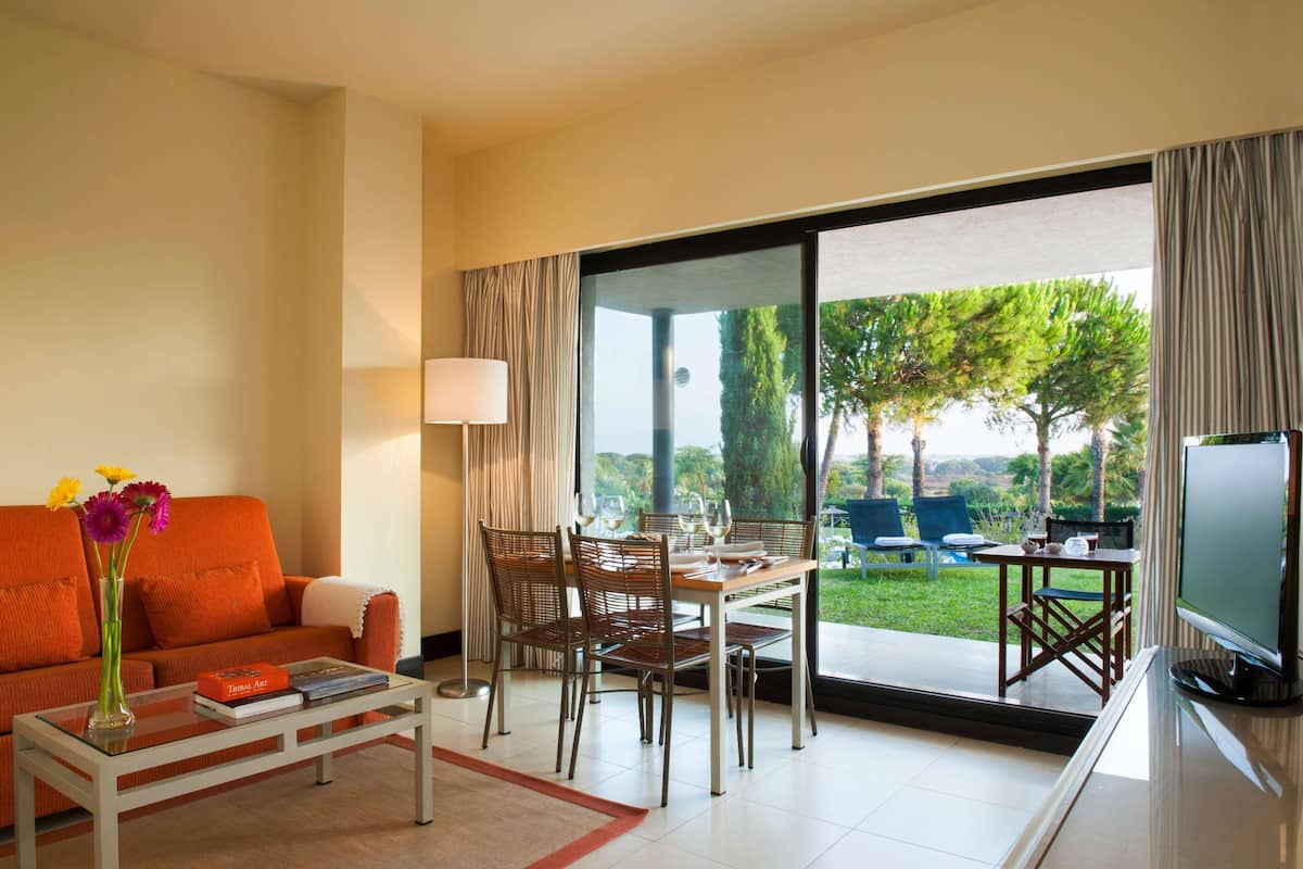 A modern living room with a sofa, coffee table, dining table, and chairs. There is a TV and a view of the outdoors through large windows.