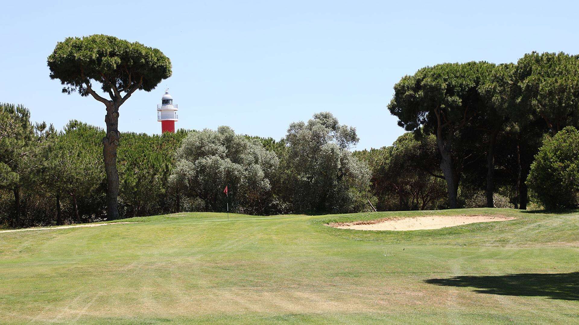 Un terrain de golf avec un phare en arrière-plan, entouré d’arbres et sous un ciel dégagé.
