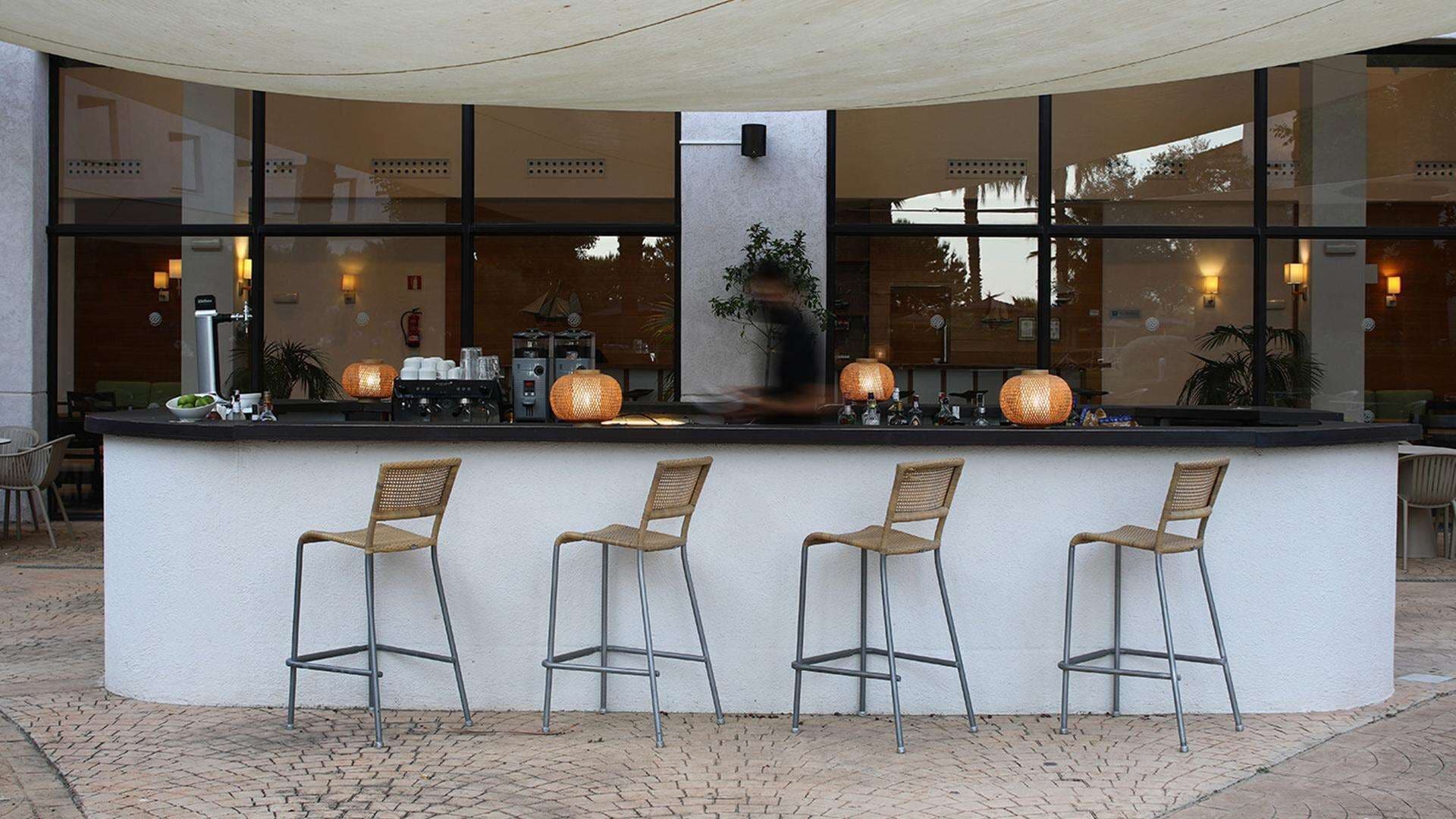 An outdoor bar area with a white counter, three wicker bar stools, and large windows reflecting the interior.